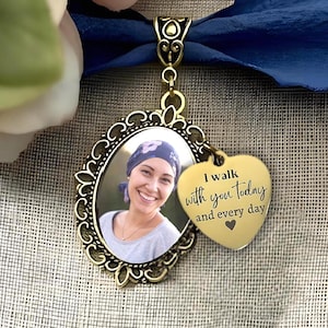 Puede incluir: Un colgante ovalado de metal en tono dorado antiguo con la fotografía de una persona sonriente que lleva un pañuelo en la cabeza. Incluye un pequeño dije en forma de corazón grabado con el texto I walk with you today and every day y un corazón.