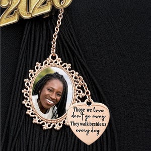May include: A graduation tassel charm with a gold-colored "2026" emblem. The charm features a photo in an ornate frame and a heart-shaped pendant with the text "Those we love don't go away. They walk beside us every day."