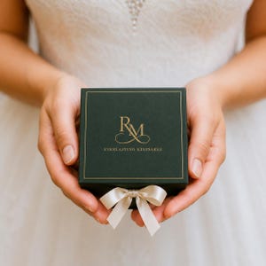 Puede incluir: Una caja de regalo cuadrada de color verde oscuro con un borde dorado y el monograma RM en color dorado, con el texto EVERLASTING KEEPSAKES impreso debajo. Un peque&ntilde;o lazo de cinta de sat&eacute;n de color claro est&aacute; sujeto en el centro inferior.