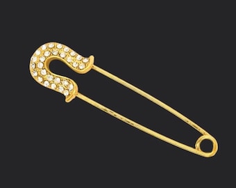 May include: A gold-colored safety pin with a curved top encrusted with small, clear crystals. The pin is set against a dark background, highlighting its shiny, metallic appearance. The pin's design is simple yet elegant, suitable for various uses.