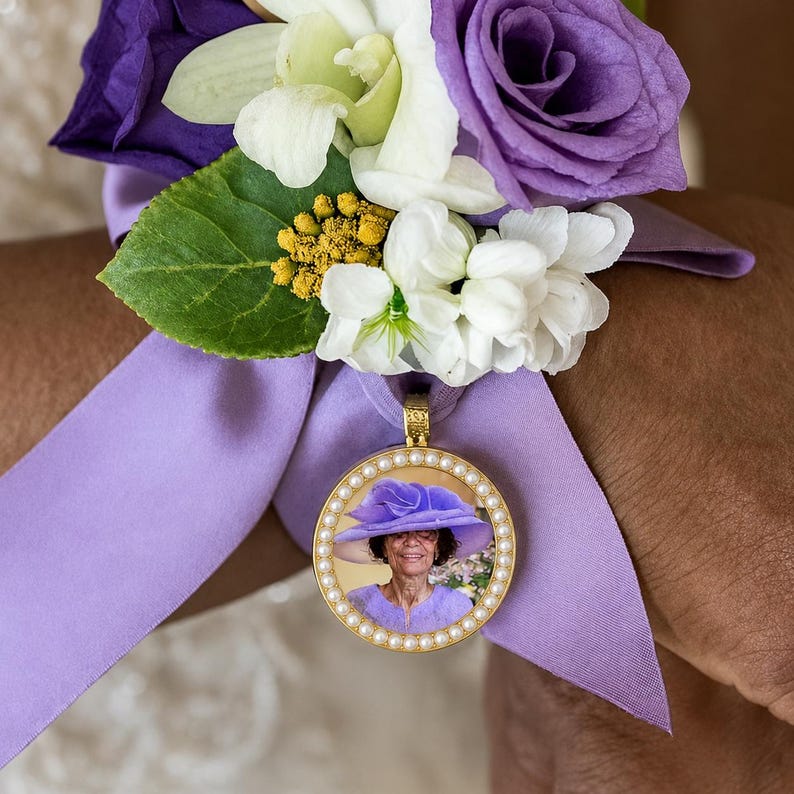 Puede incluir: Un ramillete de mu&ntilde;eca con rosas moradas, orqu&iacute;deas blancas y peque&ntilde;as flores amarillas sujeto a una cinta de sat&eacute;n morado claro. Un colgante redondo dorado con borde de perlas muestra la fotograf&iacute;a de una persona con un sombrero morado.