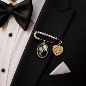Puede incluir: Un broche de imperdible en tono dorado adornado con perlas blancas, que presenta un colgante ovalado con una fotografía en blanco y negro de dos personas y un dije en forma de corazón grabado con el texto "We walk with you today and every day".