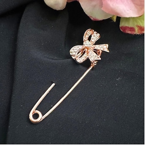 May include: A rose gold safety pin brooch with a bow design. The bow is encrusted with clear crystals. The pin is displayed on a black fabric background, with a blurred pink flower in the upper right corner.