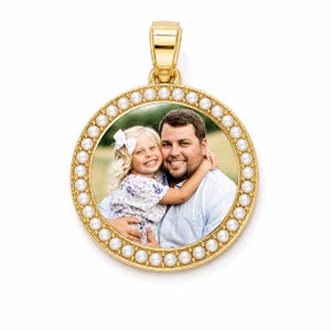 Puede incluir: Un colgante circular de oro con una fotograf&iacute;a central de un ni&ntilde;o sonriente con un lazo blanco en el pelo, sostenido por un adulto sonriente. El colgante est&aacute; enmarcado por un borde de peque&ntilde;as perlas blancas y tiene una anilla dorada.