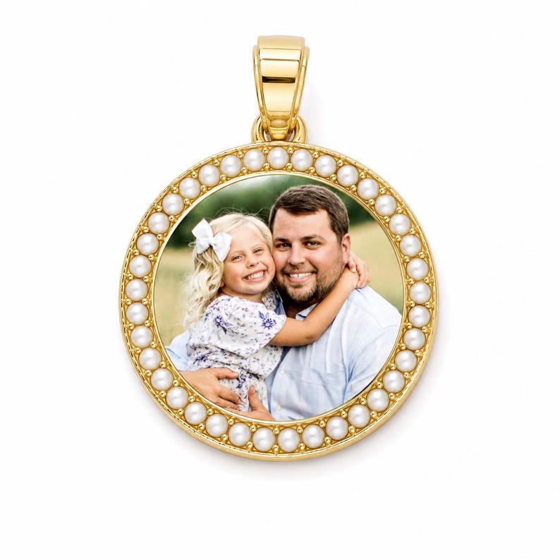 Puede incluir: Un colgante circular de oro con una fotograf&iacute;a central de un ni&ntilde;o sonriente con un lazo blanco en el pelo, sostenido por un adulto sonriente. El colgante est&aacute; enmarcado por un borde de peque&ntilde;as perlas blancas y tiene una anilla dorada.