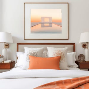 May include: A bedroom interior with a bed dressed in white bedding, decorative cushions, and an orange lumbar pillow. A framed artwork showing a minimalist structure over a pool is above the bed. Two lamps are on either side.