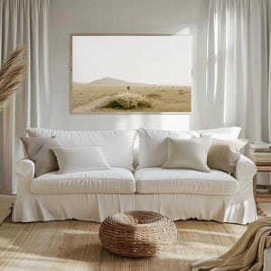 Puede incluir: Un salón con un sofá blanco y cojines de tonos neutros. Una obra de arte paisajística enmarcada cuelga sobre el sofá. Un otomán tejido y una alfombra de yute están frente al sofá. Cortinas blancas enmarcan la escena.