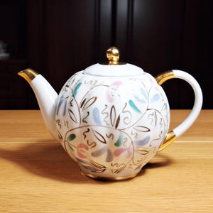 Peut inclure: Théière en céramique blanche avec une anse, un bec et un bouton de couvercle dorés. La théière est décorée d'un motif floral rose, bleu et vert, avec des accents dorés. La théière est posée sur une surface en bois.