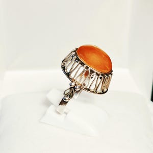 Pode incluir: Um anel de prata com uma pedra laranja grande, lisa e de formato oval, engastada numa galeria de metal decorativa e vazada. O aro inclui um pequeno detalhe em filigrana onde se une à cravação, com um acabamento metálico polido.