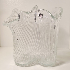Può includere: Un vaso in vetro trasparente con un bordo ondulato e una texture a coste verticali su tutto il corpo. Il vaso presenta un piccolo adesivo vicino al bordo superiore con il testo muurla hand made in finland, che ne indica l'origine e la fattura.