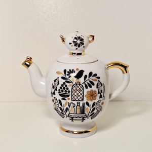 Pode incluir: Bule de cerâmica branco com detalhes dourados e um design decorativo com ilustrações pretas e douradas de pássaros, peixes e outros elementos. O bule tem uma tampa pequena com um desenho floral e uma alça dourada.