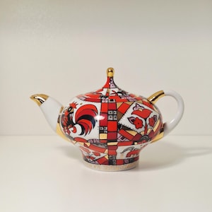 Pode incluir: Uma chaleira decorativa com um desenho de galo. A chaleira tem um bico e uma alça brancos com acabamentos dourados. O corpo é adornado com um padrão geométrico vermelho, preto e dourado e uma ilustração de galo.