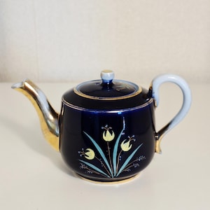 Op de afbeelding: Een donker marineblauwe keramische theepot met een goudkleurige tuit en handvat. Het lichaam is versierd met een handgeschilderd bloemmotief in lichtgeel en lichtblauw, geaccentueerd door gouden randen langs het deksel, de voet en de tuit.