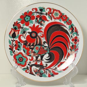 May include: A decorative white plate featuring a vibrant rooster design in red, black, and teal, surrounded by a floral border. The plate has a gold rim and is displayed on a clear stand.