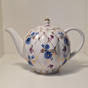 Può includere: Teiera in porcellana bianca con corpo rotondo, beccuccio curvo e manico ad anello. Decorata con disegni floreali blu e viola, dettagli dorati e un pomello dorato sul coperchio. Il design è un reticolo.