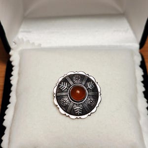 Op de afbeelding: Een ronde zilveren broche met een centrale oranje edelsteen. Het metalen oppervlak is geoxideerd met gegraveerde bloem- en pijlmotieven in een radiaal patroon. De buitenrand van de broche heeft een geschulpte, bloembladachtige afwerking.