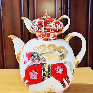 Może przedstawiać: Biały porcelanowy czajniczek z pokrywką w kształcie mniejszego czajniczka, ozdobiony żywymi czerwonymi, czarnymi i złotymi wzorami. Projekt zawiera stylizowanego konia, motywy kwiatowe oraz złote akcenty na uchwycie i wylewce naczynia.