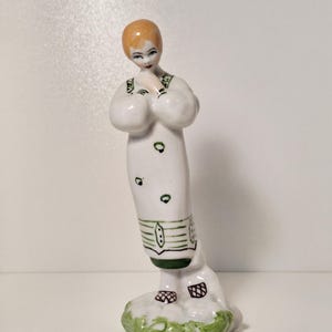 Op de afbeelding: Een porseleinen beeldje van een vrouw in een witte jurk met groene accenten. Het beeldje heeft een bobkapsel en staat op een groen-witte basis. De jurk heeft groene details en het beeldje houdt haar handen samen.