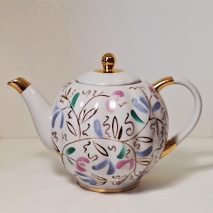 Puede incluir: Una tetera de cerámica blanca con cuerpo redondeado y acanalado que presenta un patrón floral pintado a mano en rosa, azul y verde. La tetera incluye un pico, asa y remate de tapa con bordes dorados, con detalles en oro que delinean el diseño.