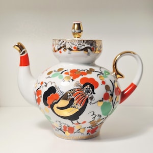 May include: White ceramic teapot with a rooster design. The teapot features a red and gold handle, spout, and lid accents. The body is decorated with a rooster, flowers, and gold detailing. A decorative piece for the kitchen or dining room.