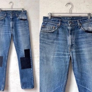 May include: Light blue denim jeans with two dark blue patches on the knees, hanging on a metal hanger. The jeans have a classic five-pocket design and a button fly. The denim has a faded, worn look.