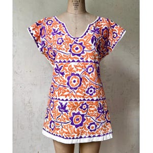 Pode incluir: Vestido túnica branco com bordados florais laranja e roxos. O vestido apresenta decote em V e mangas curtas. O bordado inclui flores, folhas e pássaros. A bainha e as bordas das mangas são finalizadas com acabamento branco.