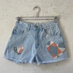 Pode incluir: Shorts de ganga azul claro com bordas desfiadas e duas aplicações de coração de patchwork. Os corações são feitos de uma variedade de tecidos em tons de azul, vermelho e branco.