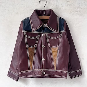 May include: A burgundy leather jacket with a contrasting navy blue yoke and tan accents. The jacket features white stitching, two chest pockets, and silver buttons. A vintage-inspired piece.