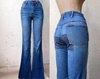 May include: High-waisted blue denim jeans with a flared leg silhouette. The back view displays a contrasting dark blue patch on the seat and red stitching details on the pockets. The denim features a medium wash with subtle fading.