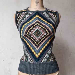Puede incluir: Un suéter sin mangas con un patrón geométrico en tonos azules, amarillos, verdes, marrones y blancos. El suéter tiene un dobladillo y cuello de canalé grises. La prenda se exhibe en un maniquí vintage.