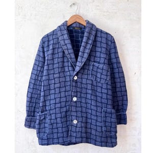 Könnte beinhalten: Blaue Jacke mit Karomuster, Schalkragen, drei weißen Knöpfen und zwei Vordertaschen. Die Jacke hängt an einem Holzbügel vor neutralem Hintergrund.