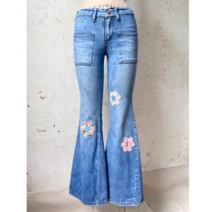 May include: Medium-wash blue denim flare jeans featuring a high-waisted fit, two front patch pockets, and three decorative floral-shaped fabric appliqués in shades of white, pink, and blue stitched onto the front of the legs.