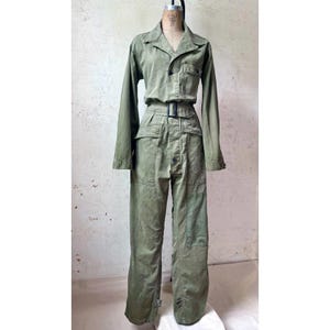 Puede incluir: Un mono de manga larga en color verde oliva con cuello, cierre frontal de botones y cinturón con hebilla rectangular. La prenda incluye dos bolsillos en el pecho, dos bolsillos en la cadera y pantalones de corte recto con diseño utilitario.