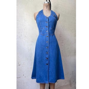 May include: A medium blue denim dress featuring a halter neckline, a fitted bodice with a button-down front closure, and a flared skirt. The garment includes a small chest pocket on the left side and visible stitching along the seams.