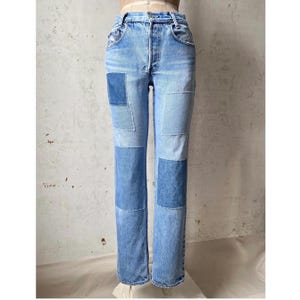 Puede incluir: Vaqueros de mezclilla azul claro con detalles de patchwork. Los vaqueros tienen un corte de pierna recta y presentan varios tonos de parches de mezclilla azul cosidos en la parte delantera de las piernas. Los vaqueros se exhiben en un maniquí.