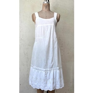 May include: A white linen dress with a square neckline and wide straps. The dress features a gathered bodice and a scalloped hemline with eyelet embroidery. The dress is displayed on a dress form.