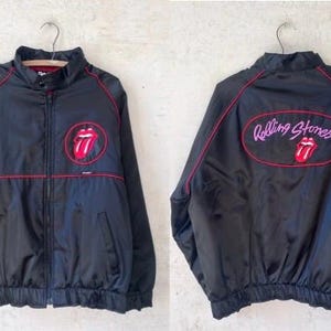 May include: Black nylon jacket with red accents and a Rolling Stones logo on the front and back. The jacket has a zipper closure, a stand-up collar, and elastic cuffs and hem. The Rolling Stones logo is a red tongue and lips design.
