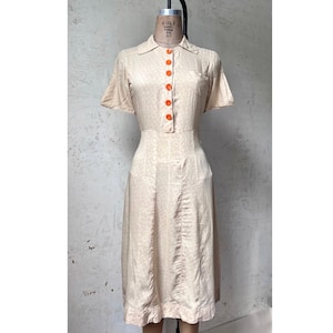 May include: A light beige midi dress featuring a subtle tonal pattern, short sleeves, and a collared neckline. The front of the dress is adorned with a vertical row of six bright orange buttons extending from the collar to the waistline.