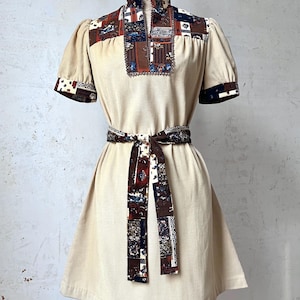 Puede incluir: Vestido beige vintage con cuello alto y mangas cortas. El vestido presenta un canesú estampado, puños y un cinturón a juego. El vestido se exhibe en un maniquí, mostrando su silueta en forma de A.