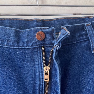 May include: Close-up of the front fly of a pair of blue denim jeans. The zipper is gold metal and the button is a brown metal button with a small design.