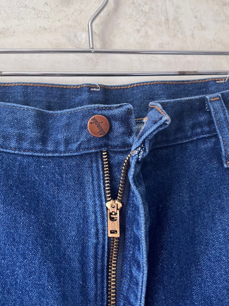 May include: Close-up of the front fly of a pair of blue denim jeans. The zipper is gold metal and the button is a brown metal button with a small design.