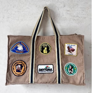 Op de afbeelding: Een bruin canvas draagtas met meerdere geborduurde patches. Patches zijn onder meer "Canadian Wilderness Voyage", "Cass Scenic Railroad", "Napa Valley Scouts", "The Pennsylvania Rangers", "Daytona International Speedway" en "Caveman Camporee". De tas heeft gestreepte handvatten.