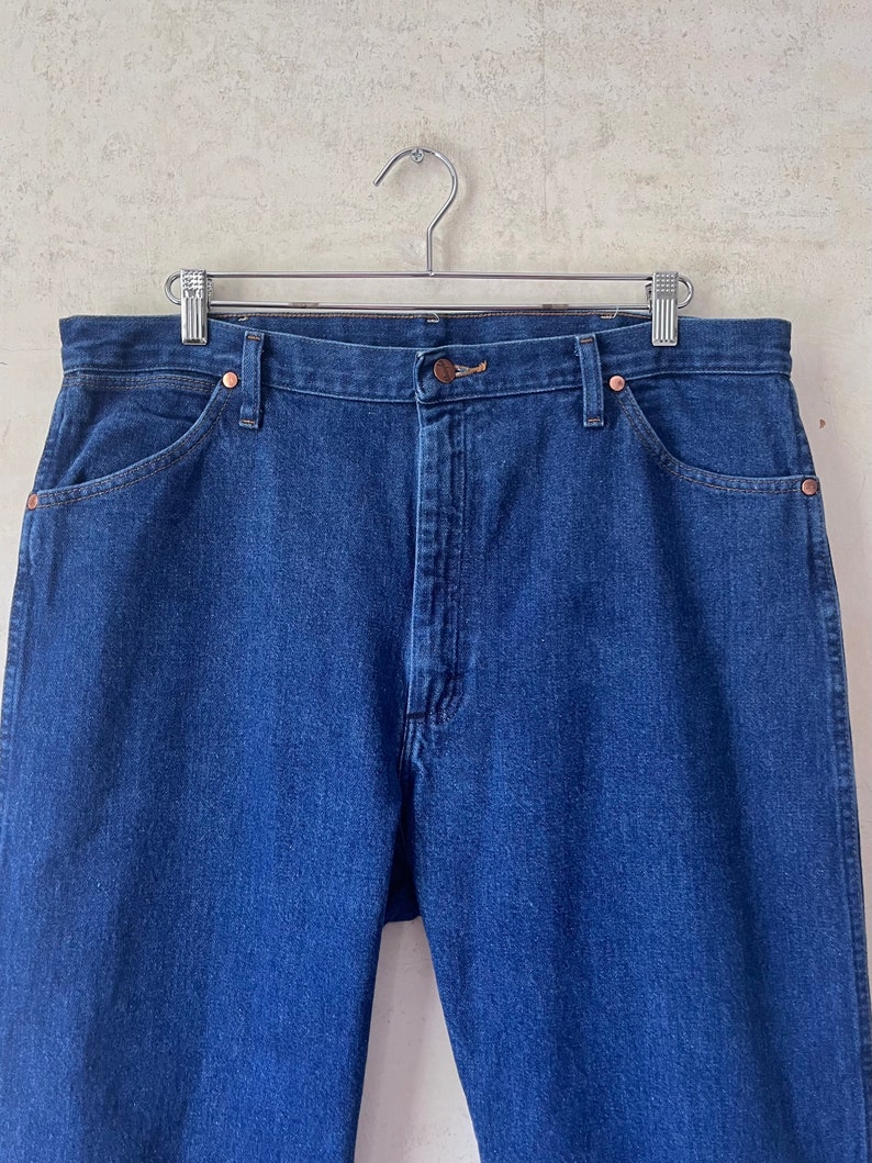 May include: A pair of dark blue denim jeans hanging on a silver hanger. The jeans have a classic five-pocket style and a straight leg.