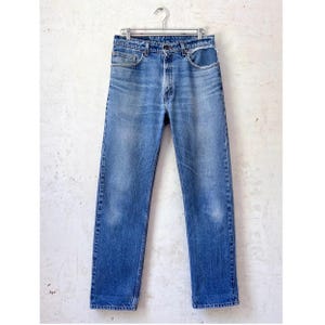 Peut inclure: Un jean en denim bleu délavé moyen avec une coupe droite classique à cinq poches et une braguette à boutons. Le tissu présente des décolorations naturelles et des marques d'usure sur le devant des cuisses et au niveau de la taille.