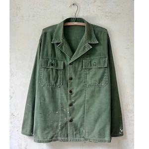 Puede incluir: Chaqueta verde oliva con botones, dos bolsillos en el pecho y dos bolsillos inferiores. La chaqueta tiene cuello y mangas largas. La tela muestra signos de desgaste, con algunas salpicaduras de pintura. La chaqueta está colgada de una percha metálica.
