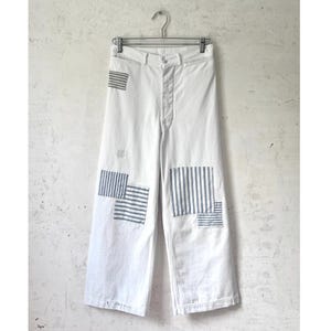 Puede incluir: Pantalones blancos de pierna ancha con cierre de botones y múltiples parches rectangulares con un patrón de rayas verticales azules y blancas. Los parches están cosidos en la parte delantera de las piernas en varios tamaños y posiciones.