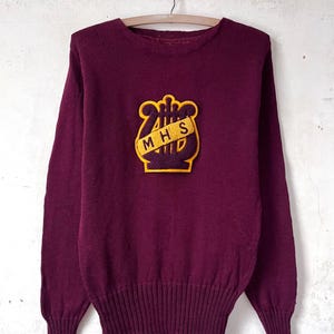 Peut inclure: Un pull maron avec un écusson brodé jaune et marron représentant une lyre et les lettres "MHS".