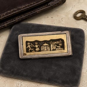 May include: A rectangular metal money clip featuring a two-tone design with a silver-colored frame and a gold-colored center inlay. The inlay displays a dark, recessed relief scene depicting two figures, a small structure, and a covered stall.