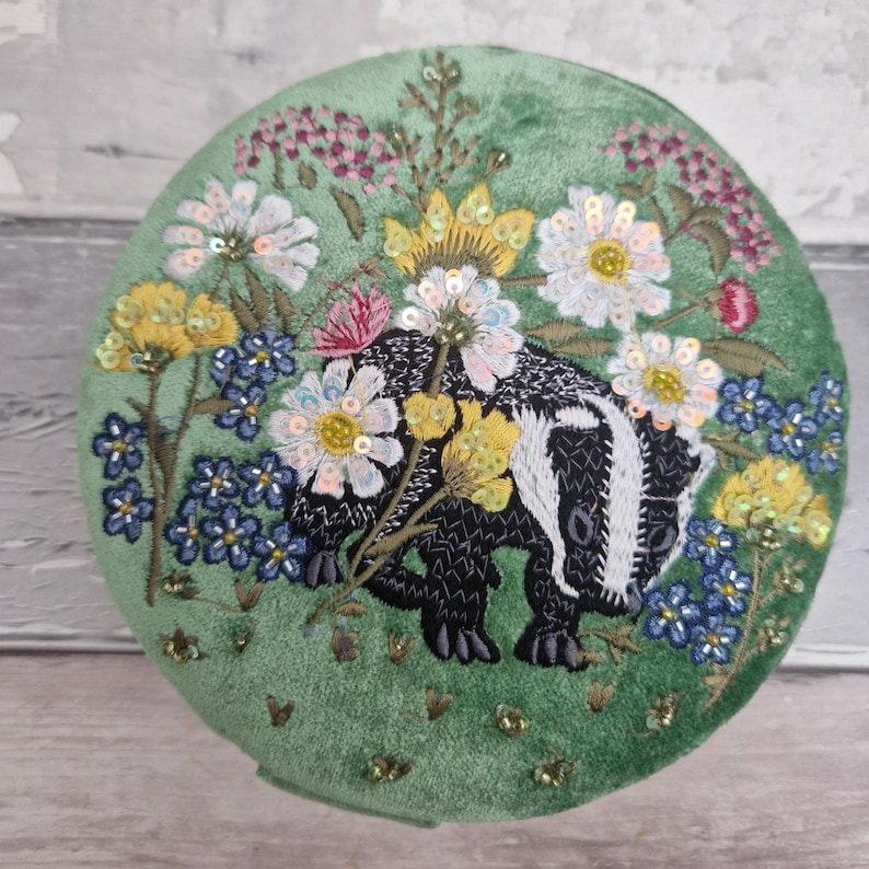 May include: A round, green velvet decorative piece featuring an embroidered badger surrounded by colorful flowers. The badger is black and white, and the flowers include white, yellow, pink, and blue blooms, with sequin accents.