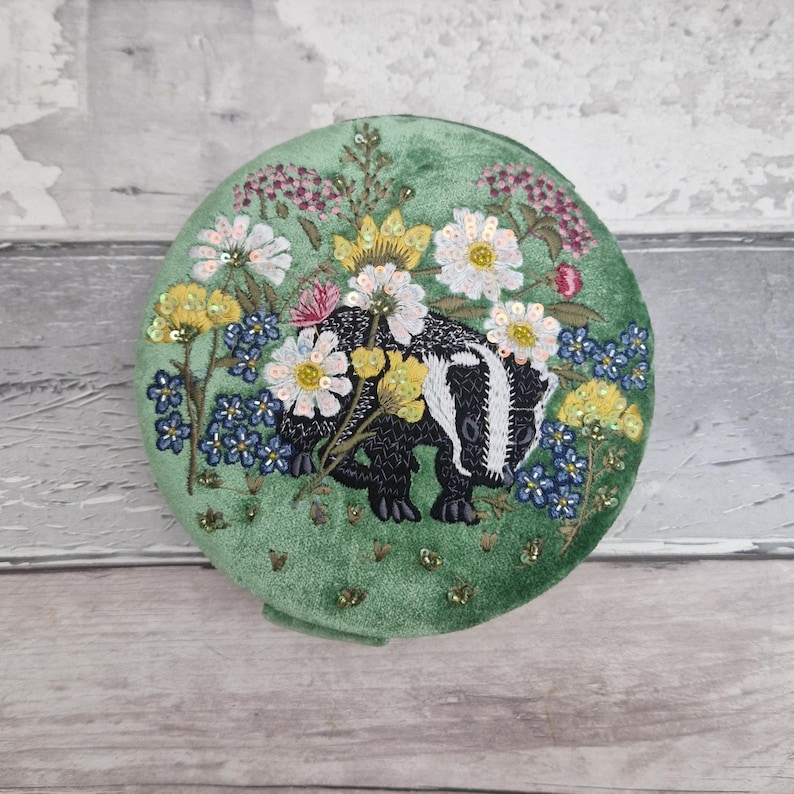 May include: Round, green velvet compact with an embroidered badger design. The badger is black and white, surrounded by colorful flowers and sequins. The compact is a unique accessory.